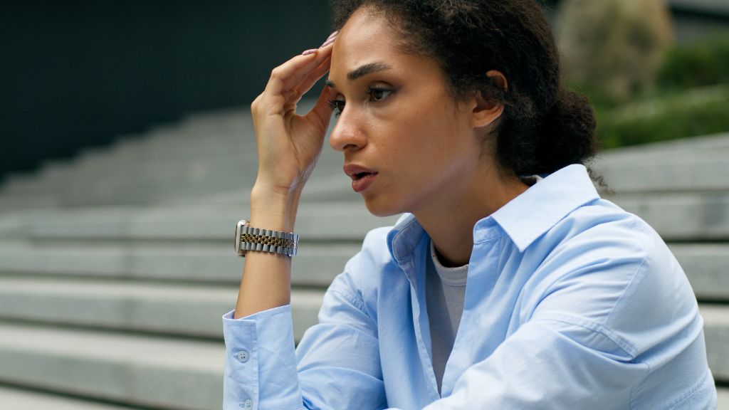 Mulher negra sentada em escadaria cinza ao ar livre, com expressão de preocupação, apoiando a mão na cabeça. Usa camisa azul-clara, relógio metálico no pulso esquerdo e cabelos presos, ilustrando saúde mental docente, professora preocupada e bem-estar escolar.