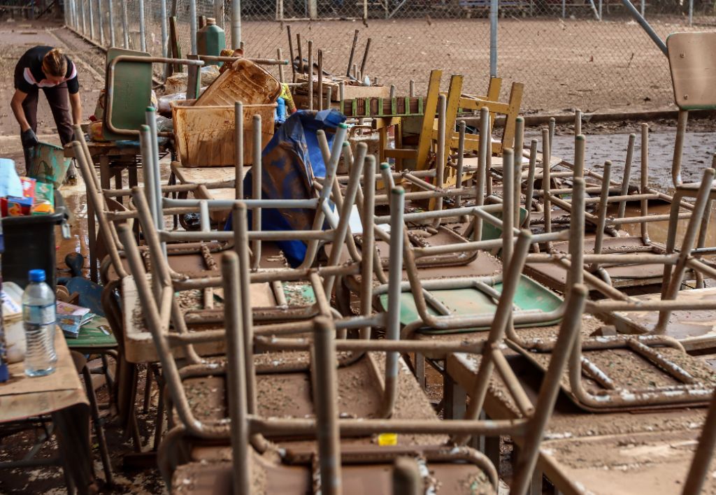 Carteiras e móveis escolares cobertos de lama após enchentes em escola no Rio Grande do Sul em 2024. Cena mostra destruição escolar, impacto das enchentes no RS em 2024 e trauma escolar, destacando os desafios da recuperação da educação. Ao fundo, uma pessoa limpando a lama com um balde.