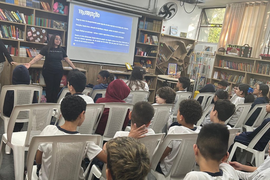 Mulher apresenta uma palestra sobre frustração para uma turma de alunos em uma biblioteca escolar.